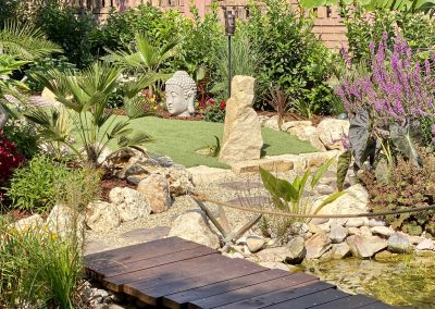 A kerti tó felett fahíd vezet át, kenderkötél korláttal, a háttérben könyöklőszikla.